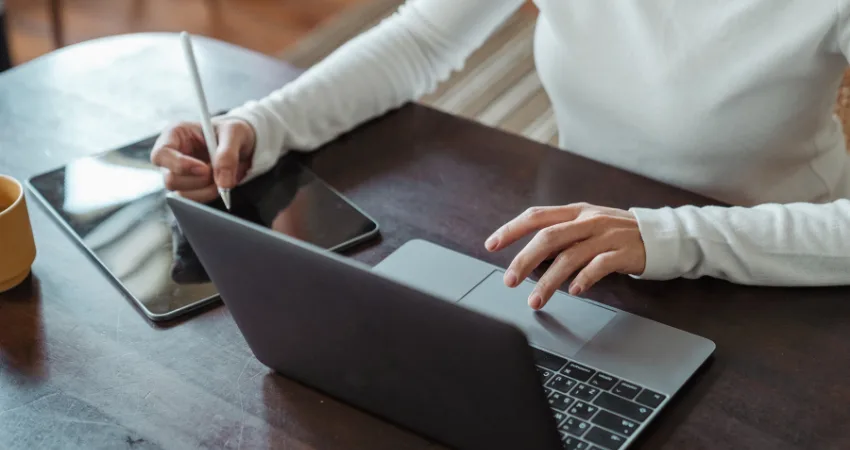 شخص يعمل لدى شركة إيكوواي الأردن على طاولة خشبية داكنة، يستخدم جهاز كمبيوتر محمول بيد وقلمًا على جهاز لوحي باليد الأخرى. ويظهر كوب أصفر بجانب الجهاز اللوحي.