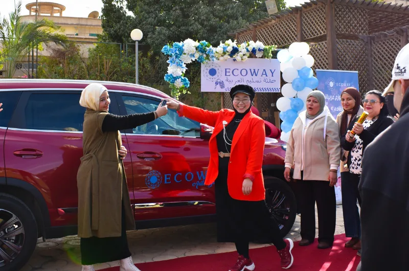 تقف امرأتان بجانب سيارة دفع رباعي حمراء على سجادة حمراء، تُسلّم إحداهما المفاتيح للأخرى. تحمل اللافتات عبارة "تقف امرأتان بجانب سيارة دفع رباعي حمراء على سجادة حمراء، تُسلّم إحداهما المفاتيح للأخرى. تحمل اللافتات عبارة ECOWAY وحاملة توصيل العربية، كما تحمل السيارة أيضاً عبارة ECOWAY وتوصيل