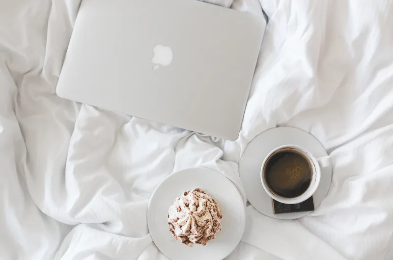 An overhead view displays a silver laptop, a coffee cup, and a dessert on a plate, all resting on white, rumpled bedsheets, suggesting a relaxed morning.