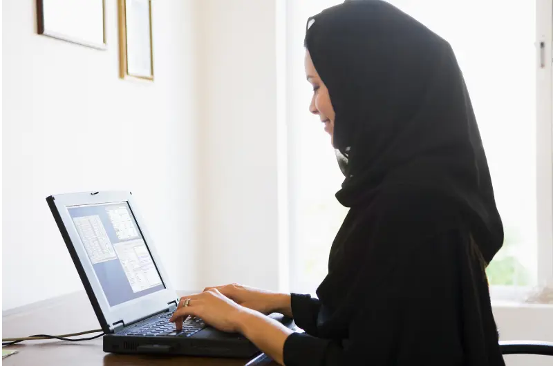 امرأة ترتدي حجاباً أسود تبتسم برفق أثناء الكتابة على جهاز كمبيوتر محمول، كما يظهر من الجانب. تعرض شاشة الكمبيوتر المحمول أعمدة من النصوص.