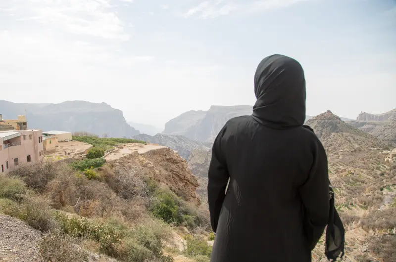 امرأة ترتدي عباءة سوداء تقف وظهرها للمشاهد، تحدق في منظر طبيعي  بالاردن جبلي شاسع وجاف مع بعض المباني على اليسار.
